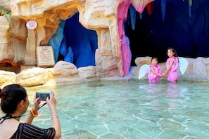 A pool with clear water, a rocky cave-like structure, and two children in pink dresses.