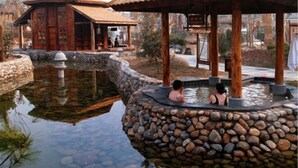 Una estructura de madera tradicional sobre base de piedra, una fuente de piedra y una pequeña piscina donde la gente se relaja.