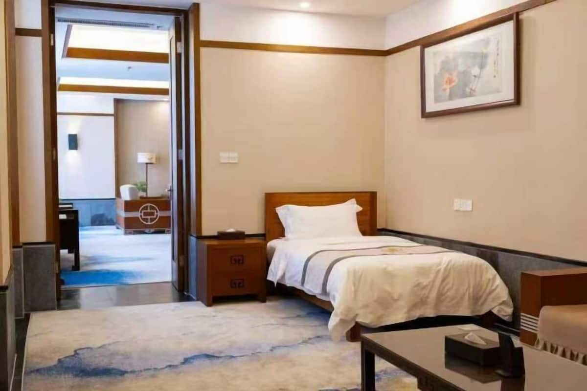 A hotel room with a bed, bedside table, and a desk. There is a door leading to another room and a window with a view.