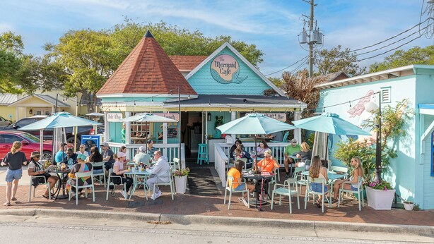 A seaside cafe with outdoor seating, a red roof, and a sign reading 'The Mermaid Cafe.'