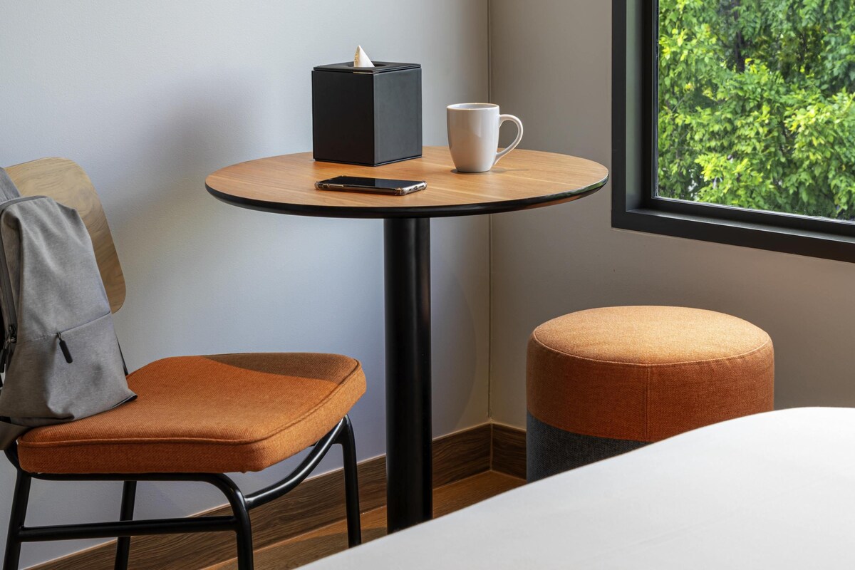 Une chambre d’hôtel avec une table ronde en bois, une tasse blanche, un smartphone et un objet noir posé sur la table.