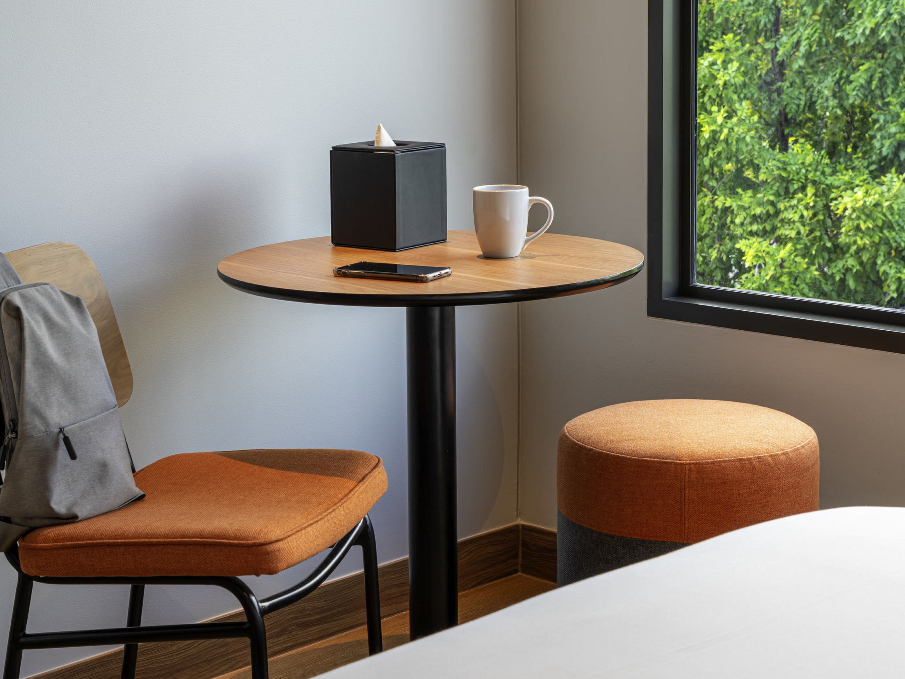 Una habitación de hotel con una mesa redonda de madera, una taza blanca, un teléfono inteligente y una caja de pañuelos de papel negros.