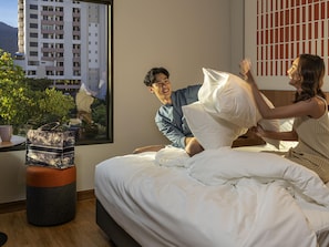 Une chambre à coucher avec un lit, une fenêtre donnant sur un immeuble, et une petite table sur laquelle se trouvent une plante et un sac.