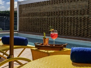 Un espace aménagé au bord de la piscine, avec des fauteuils en osier jaune, une serviette bleue et un verre d’une boisson rouge décorée d’une tranche de citron.
