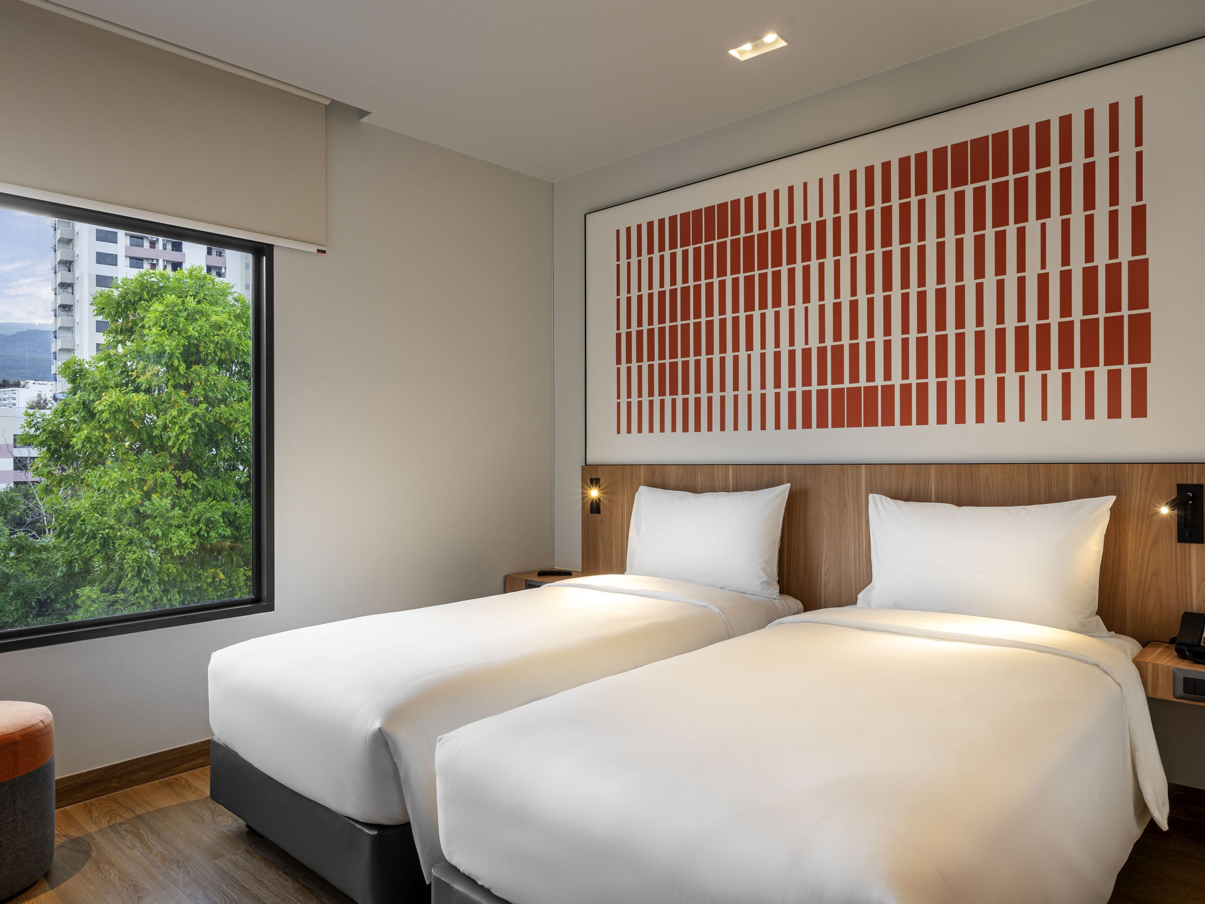 A hotel room with two beds, a large window with a view of a tree and buildings, and a decorative wall panel.