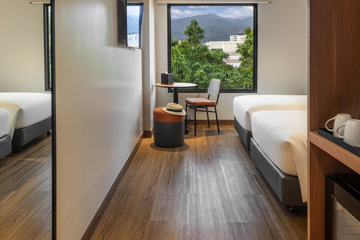 Habitación de hotel con dos camas, una mesa pequeña con una silla, un cesto de basura y una ventana con vistas a montañas y edificios.