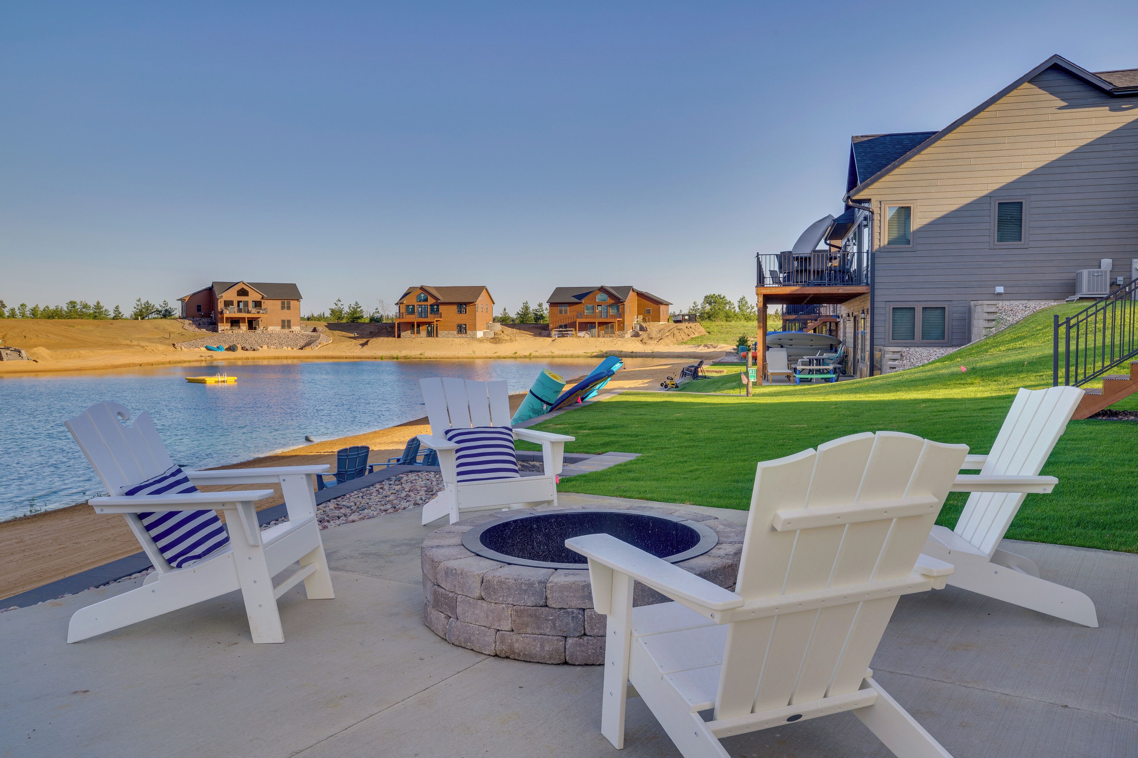 A waterfront property with a stone fire pit, white Adirondack chairs, and a view of a lake.