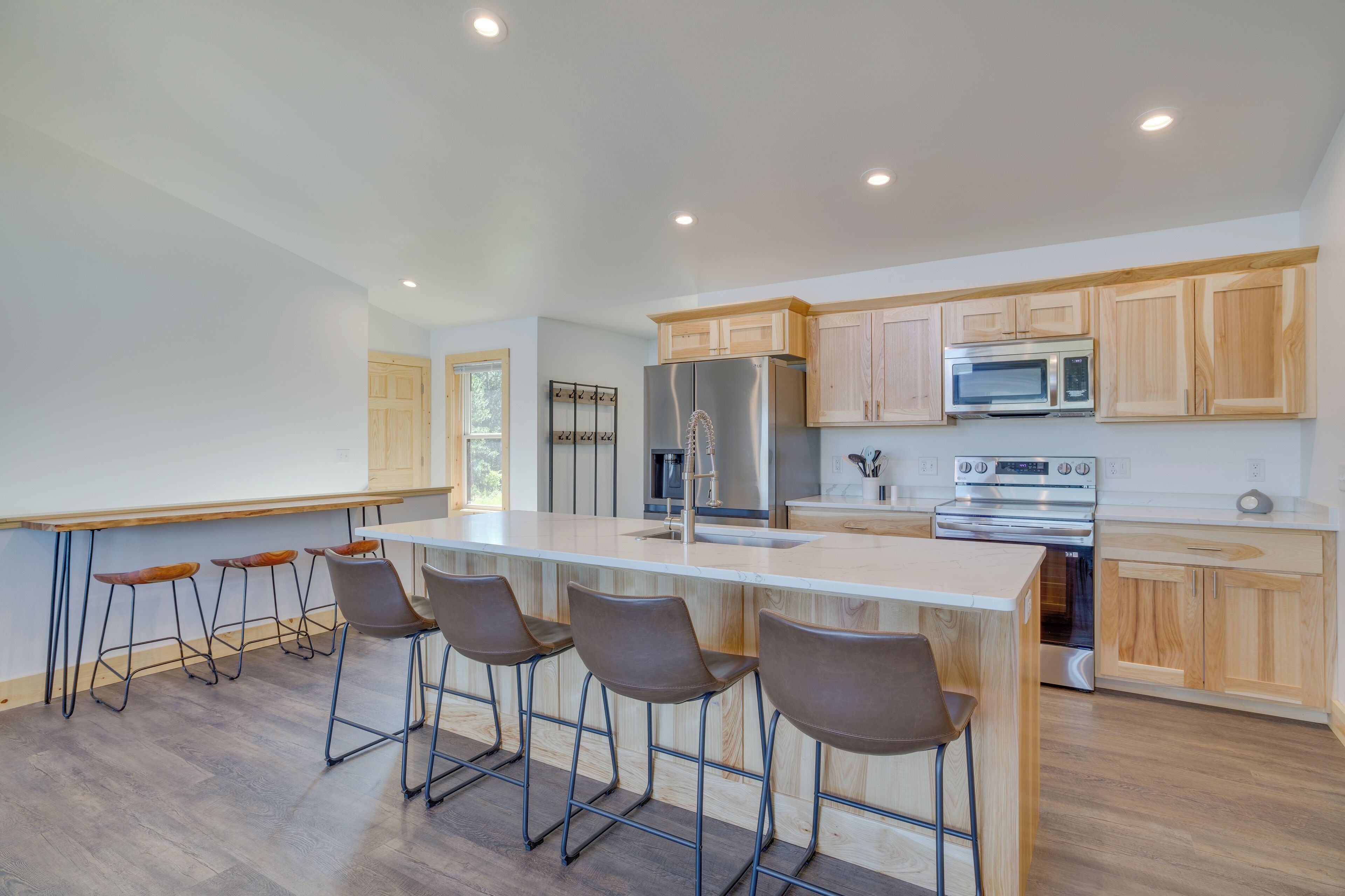 A modern kitchen with a central island, bar stools, wooden cabinets, stainless steel appliances, and a large window.