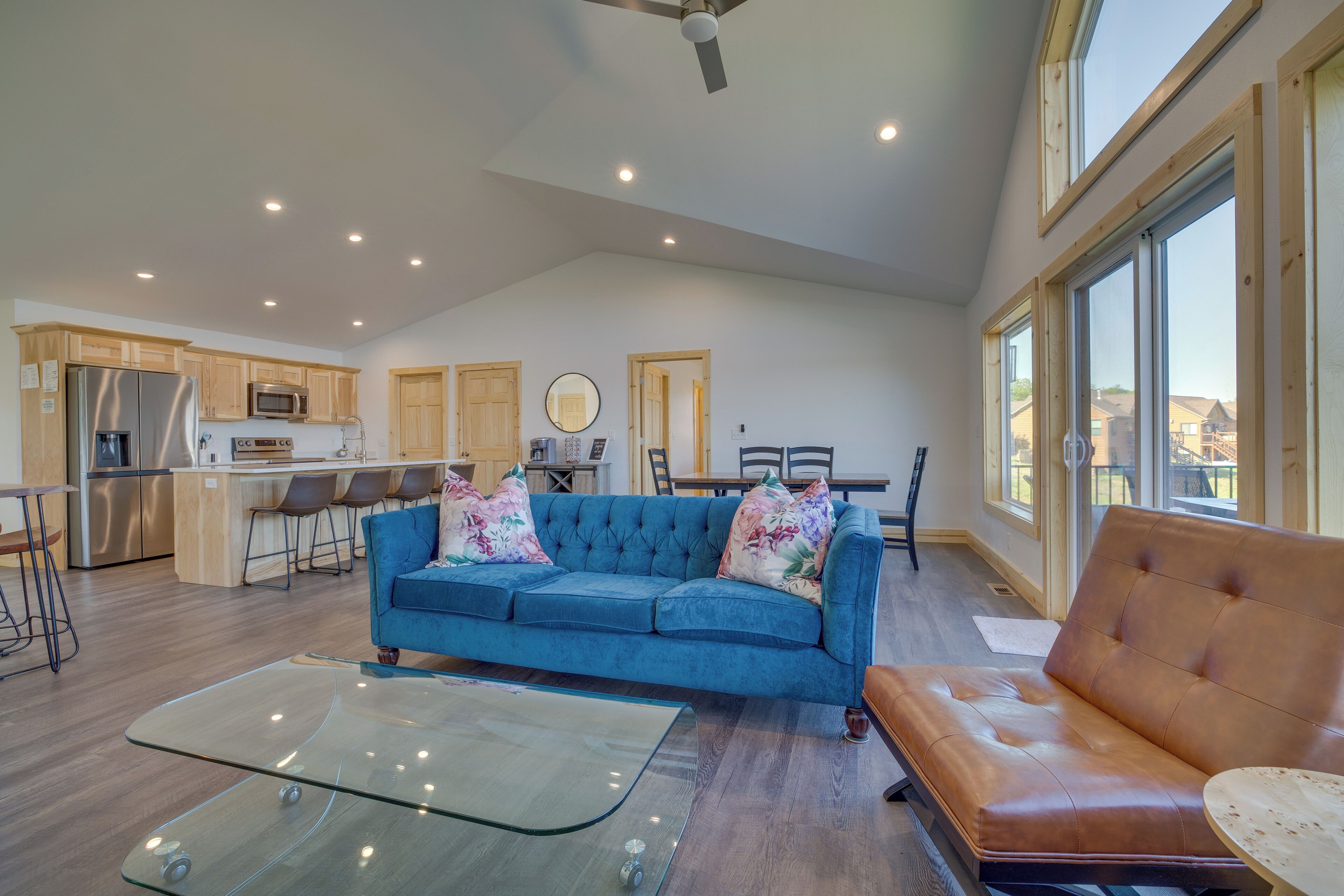 A spacious living area with a blue sofa, a brown leather couch, and a glass coffee table. There is a kitchen area with wooden cabinets and a stainless steel refrigerator.