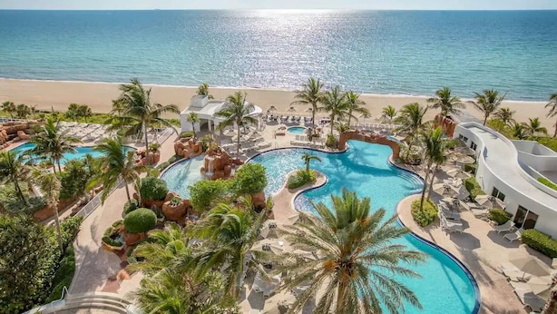 Un resort con una amplia piscina central, palmeras y vista a la playa.