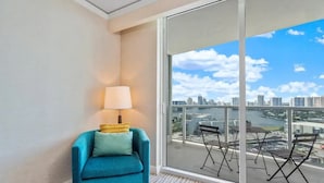 Habitación de esquina con un sillón color turquesa, una lámpara y vistas a un paisaje urbano a través de amplios ventanales.