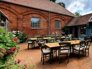 A brick building with a tiled roof, outdoor seating area with wooden tables and chairs, and a garden with roses.