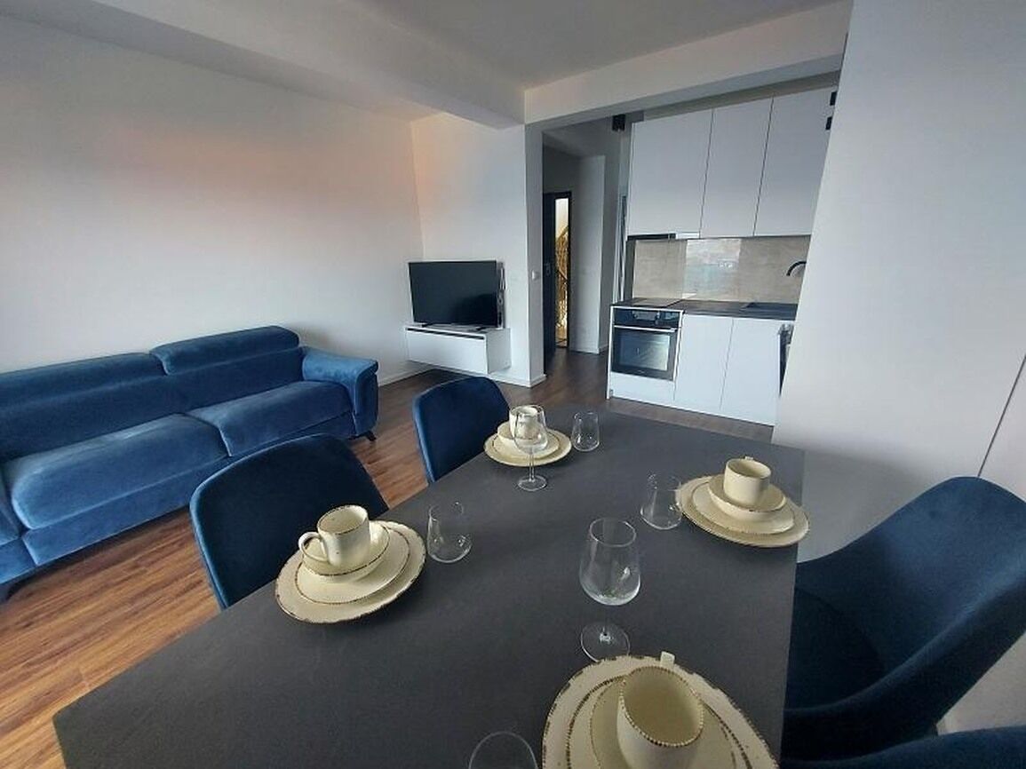 Ein modernes Wohnzimmer mit einem blauen Sofa, einem Esstisch gedeckt mit Tellern und Gläsern, und einer Küche im Hintergrund.