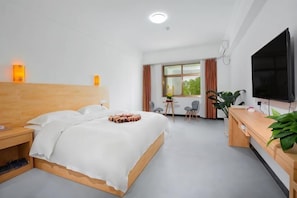 A hotel room with a wooden bed, a desk with a TV, a chair, a window with curtains, and a plant.