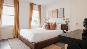 Un dormitori amb un llit, un escriptori i una cadira. Hi ha cortines a les finestres, un ventilador de sostre i làmpades penjades a la paret.