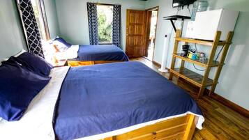 Una habitación con dos camas de madera, un estante de madera con objetos, una puerta y un televisor en la pared.