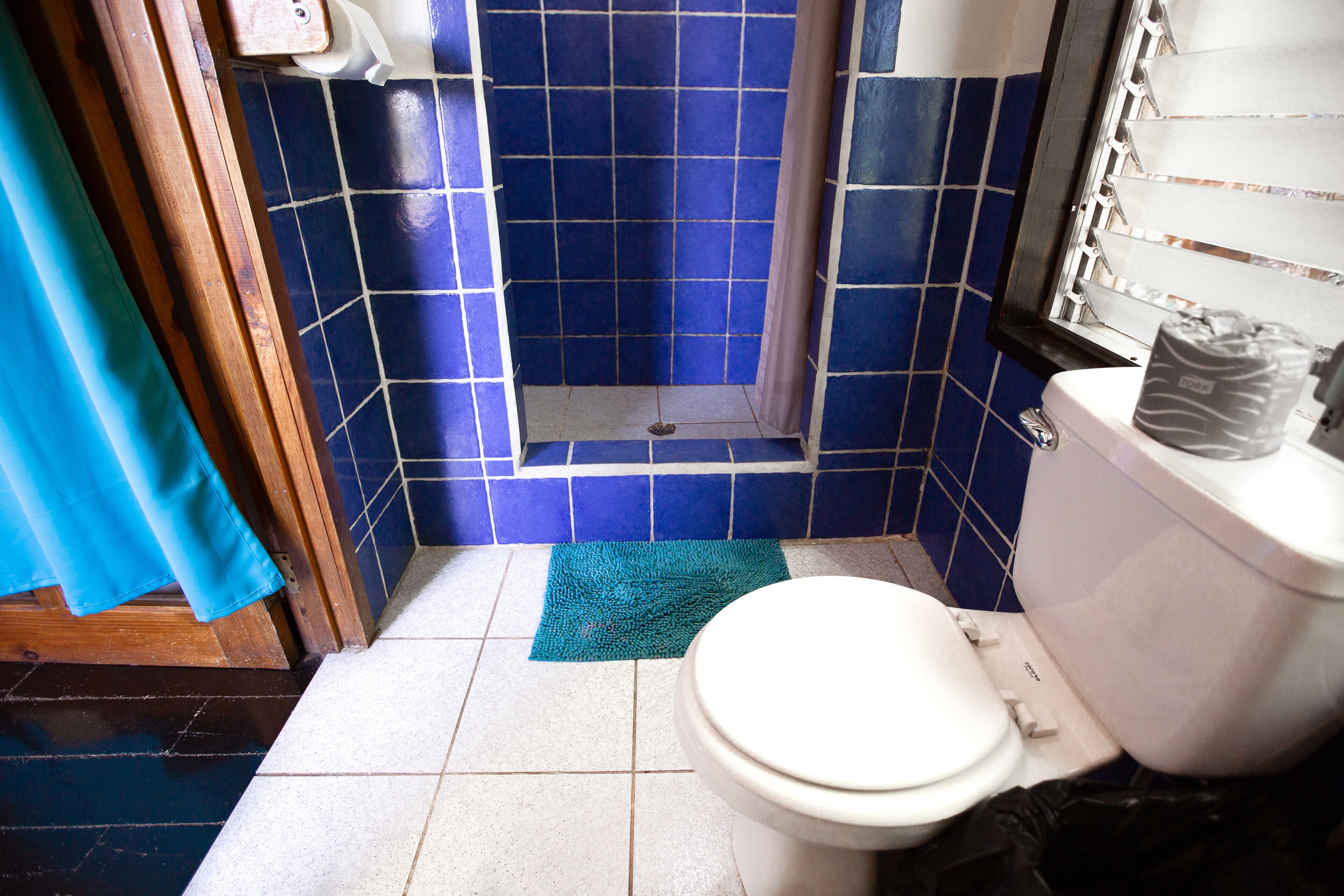 Un baño con inodoro blanco, paredes de azulejos azules, una zona de ducha y un tapete azul en el suelo.