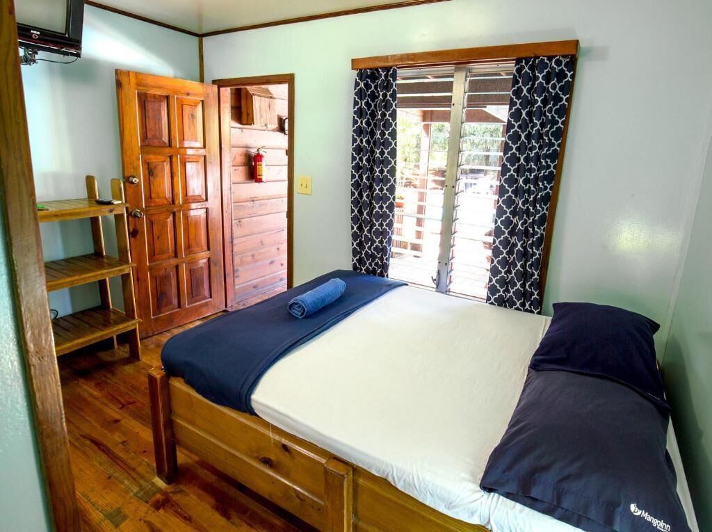 Un dormitorio con cama de madera, manta azul, almohada oscura, armario de madera y ventana con cortinas estampadas.