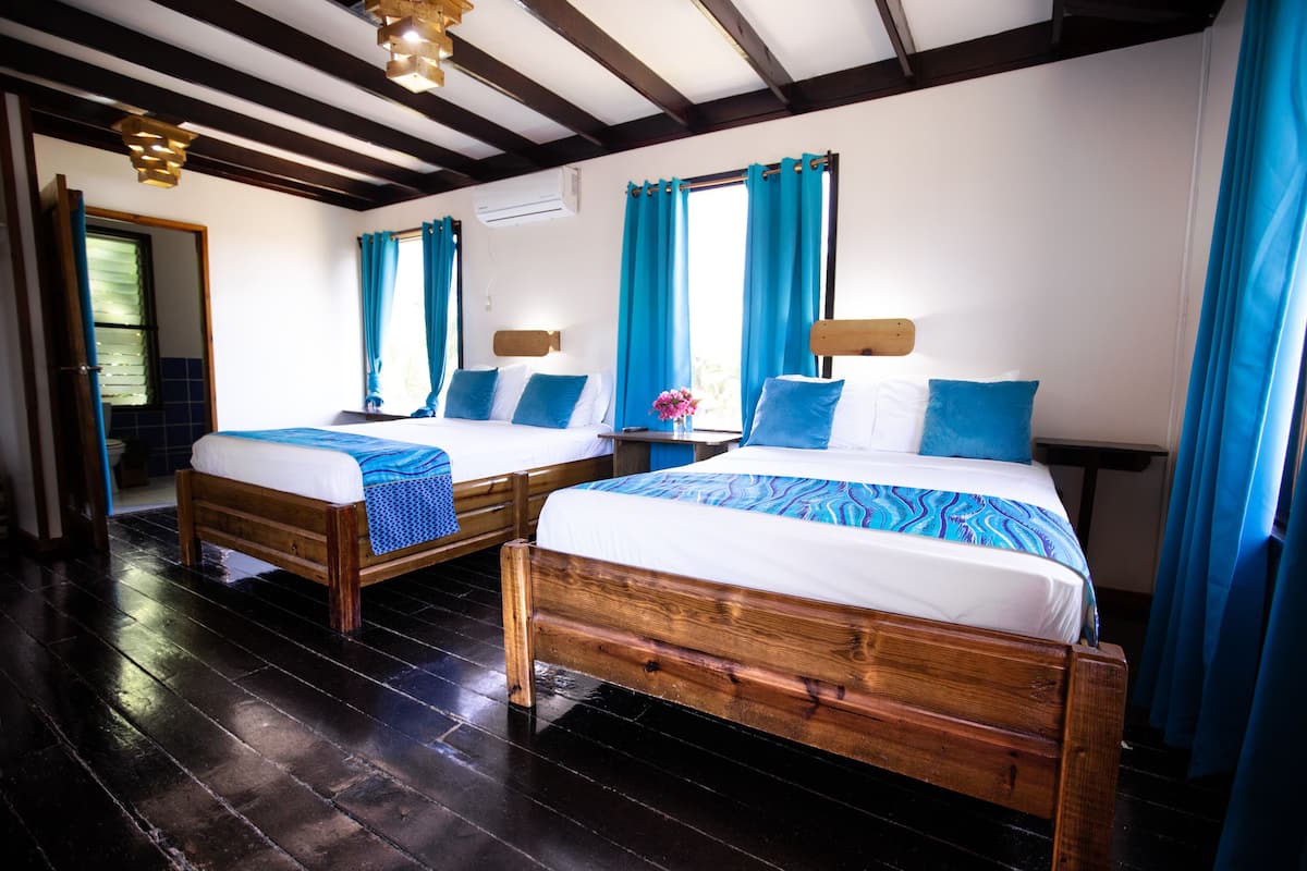 A room with two wooden beds, each with a blue and white bedspread, and a wooden headboard. The room has a dark wooden floor and a sloped ceiling with exposed beams.