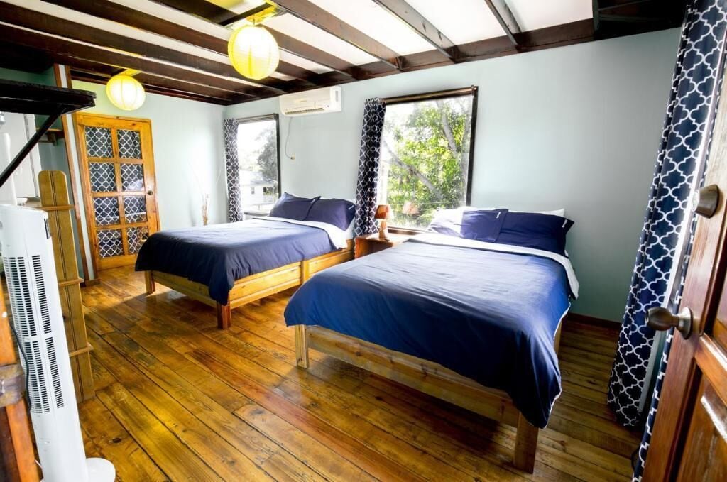 Una habitación con dos camas, suelo de madera, ventilador de techo y ventana con vista a árboles.