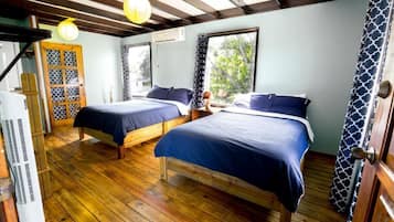 Una habitación con dos camas, suelo de madera, ventilador de techo y ventana con vista a árboles.