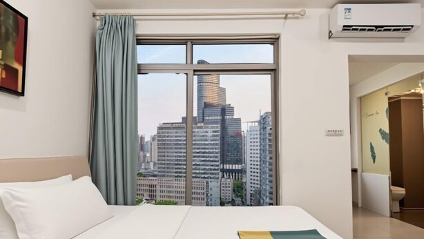 Ein Hotelzimmer mit großem Fenster, einem Bett mit weißer Bettwäsche, Blick auf Stadtgebäude und einer Klimaanlage.