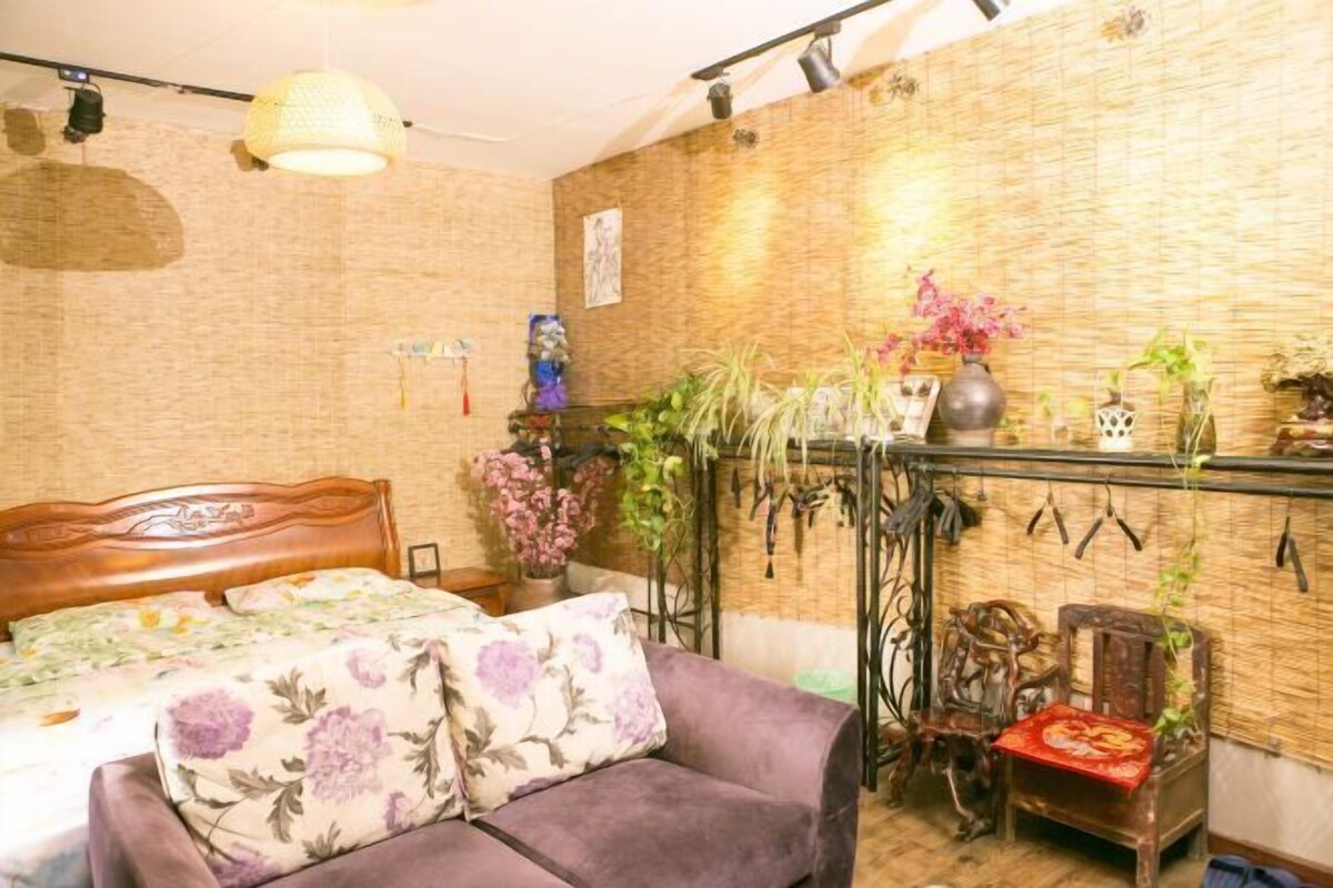 A room with a bed, a purple sofa, a wooden chair, and a wall with plants and a hanging light fixture.
