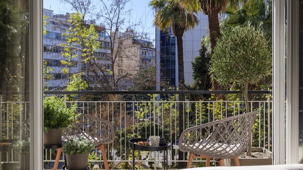 Un balcone con due sedie in vimini, un tavolino con una tazza e una pianta, e vista su edifici e alberi.