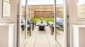 Eine Terrasse mit Tisch und Stühlen, ein kleiner Tisch mit Blumen in einer Vase sowie Blick auf einen Garten mit Holzzaun und Steinmauer.