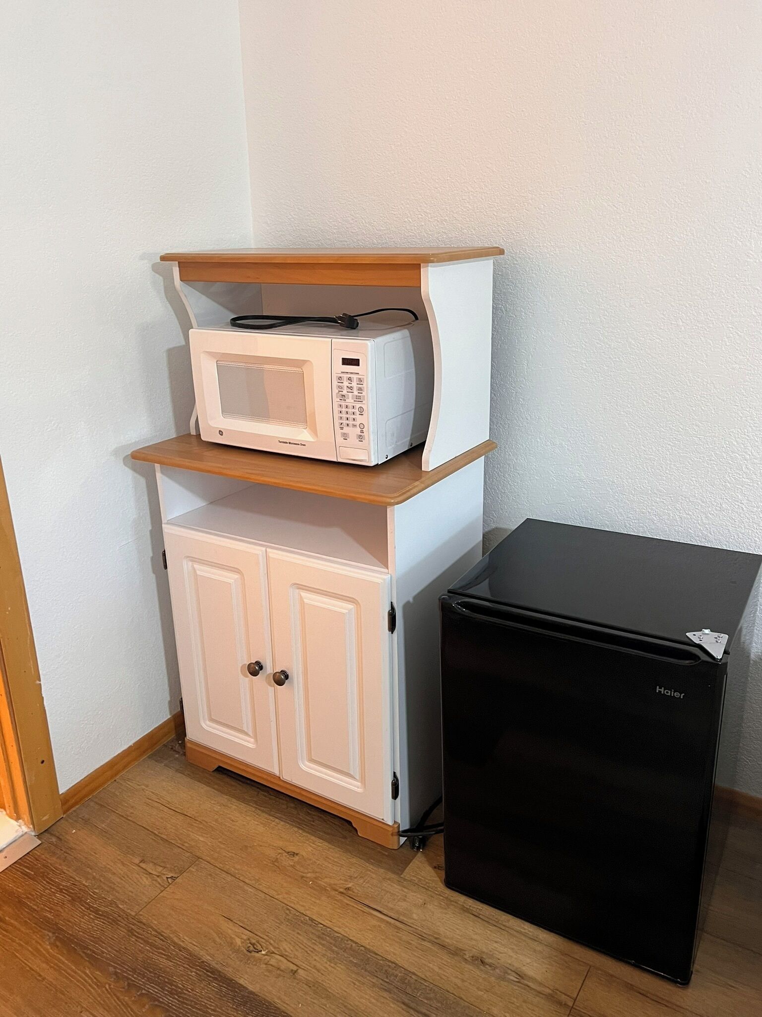 A compact kitchen area with a white microwave on a shelf, a white cabinet below, and a black refrigerator to the right.