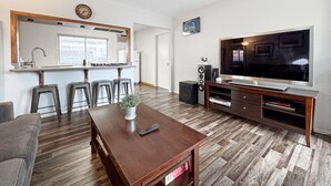 Un salon moderne avec une table basse en bois, un téléviseur posé sur un meuble en bois et un coin cuisine avec une fenêtre.