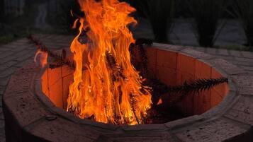 Ein kreisrunder Feuerkorb mit Flammen, umgeben von einer Ziegelumrandung, in einem Außenbereich mit Bäumen und blauem Himmel im Hintergrund.