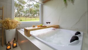 Ein modernes Badezimmer mit einer großen Badewanne, einem Holzregal mit Snacks und Getränken, einer Topfpflanze und Blick auf eine grüne Landschaft durch ein Fenster.