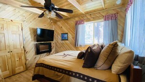 Un dormitorio tipo cabaña de madera con cama, ventilador de techo, televisor y ventanas con cortinas.