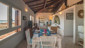A dining area with a table set for four, surrounded by white wicker chairs. There is a television on a stand to the left, and a shelf with various items above it. - Il Vecchio Faro Attico A5 (Loiri Porto San Paolo)
