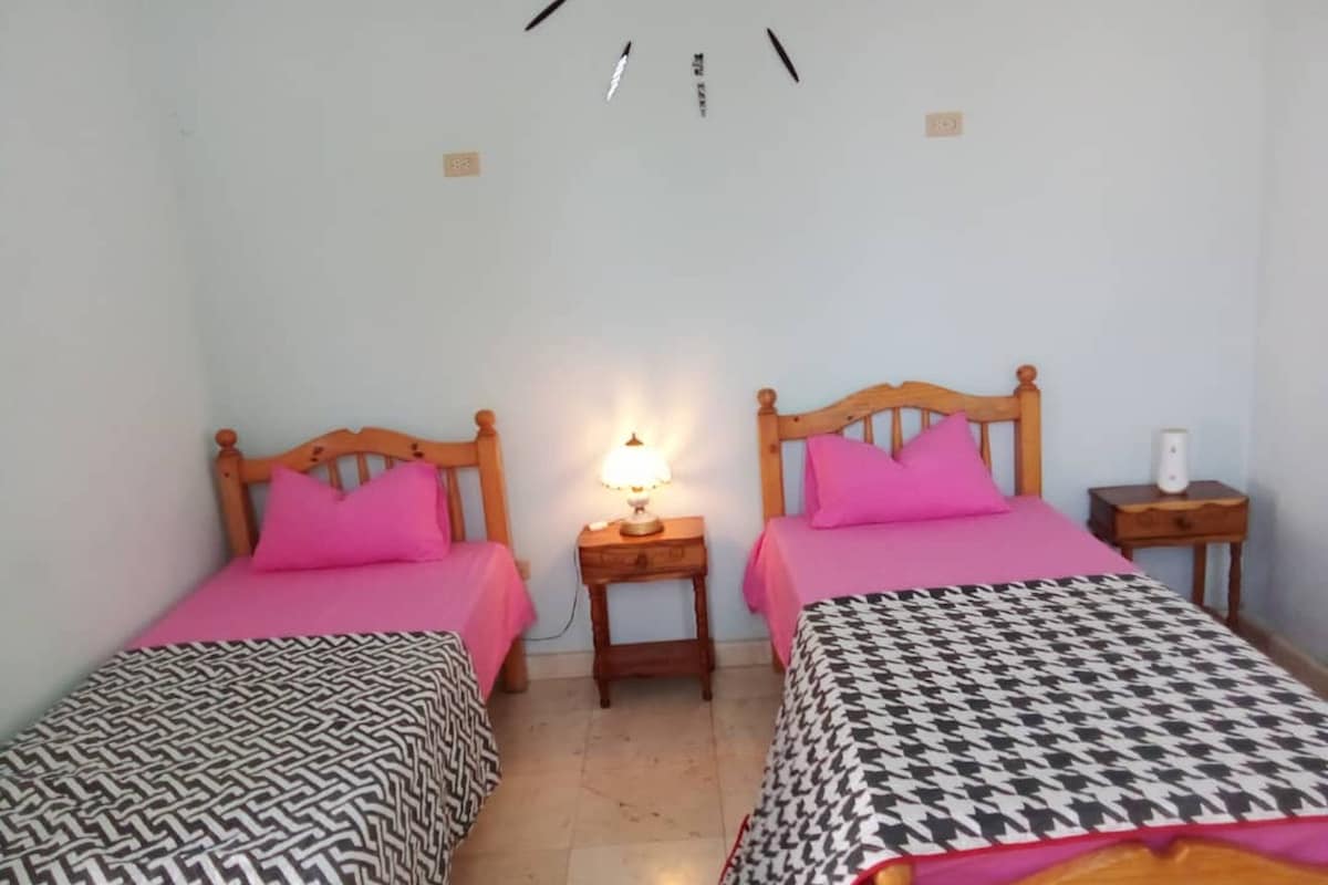 Une petite chambre propre, avec deux lits en bois, chacun agrémenté d’un oreiller rose et d’une couvre-lit à motifs. Une table de chevet avec une lampe se trouve entre les deux lits.