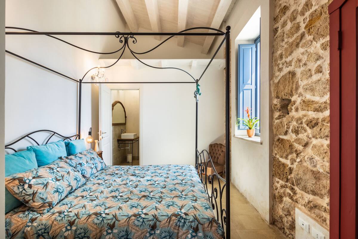 A bedroom with a four-poster bed, a stone wall, and a window with a plant.