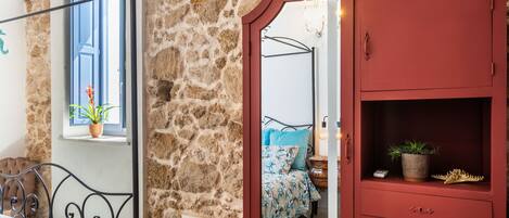 A bedroom with a stone wall, a red wardrobe, a bed with a floral bedspread, a window with a plant, and a mirror reflecting the room.