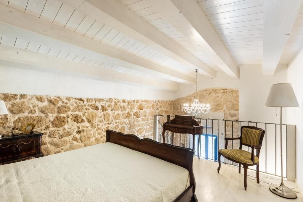 A bedroom with a stone wall, a bed, a piano, a chair, and a balcony with a railing.