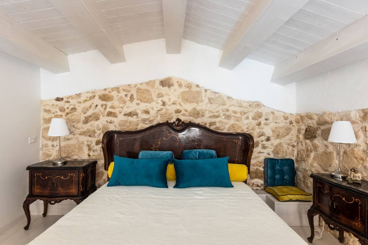 A bedroom with a stone wall, wooden ceiling beams, a bed with blue and yellow pillows, and two nightstands with lamps.