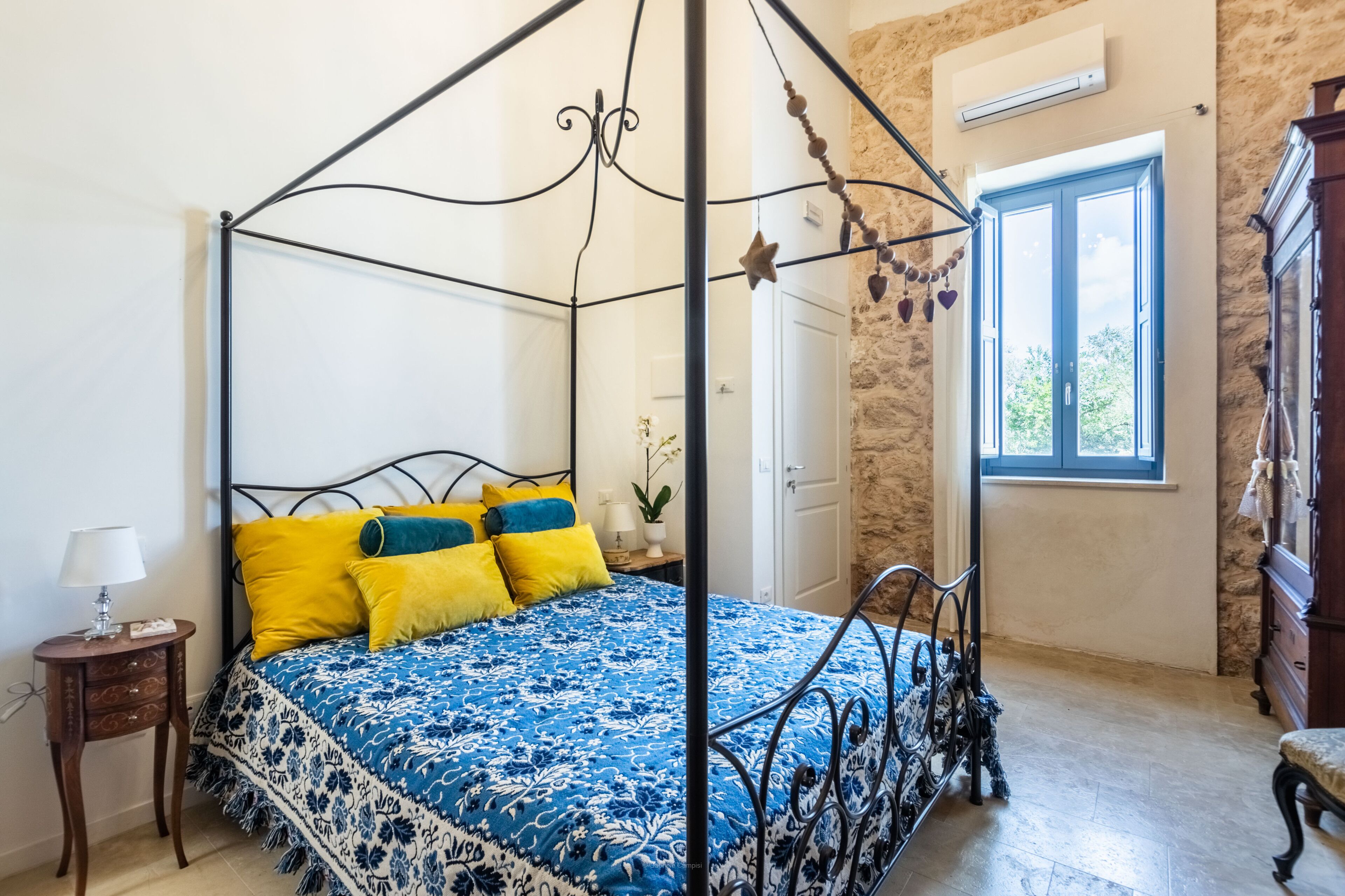 A four-poster bed with a blue floral bedspread and yellow pillows, a small bedside table with a lamp, and a stone wall with a window.