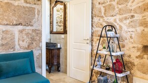 A room with a stone wall, a white door, a blue sofa, a wooden cabinet, and a metal shelf with various items. - Michael Garden (Noto)