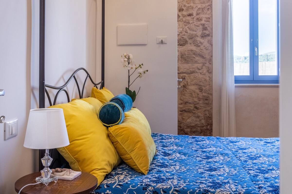 A bedroom with a metal bed frame, a blue patterned bedspread, yellow and blue pillows, a bedside table with a lamp, and a window with a star decoration.