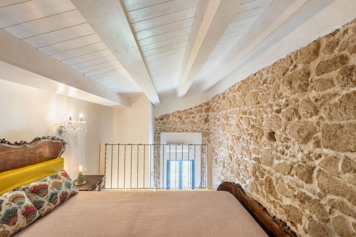 A bedroom with a stone wall, a wooden ceiling, a bed with a colorful pillow, a nightstand, and a chandelier.