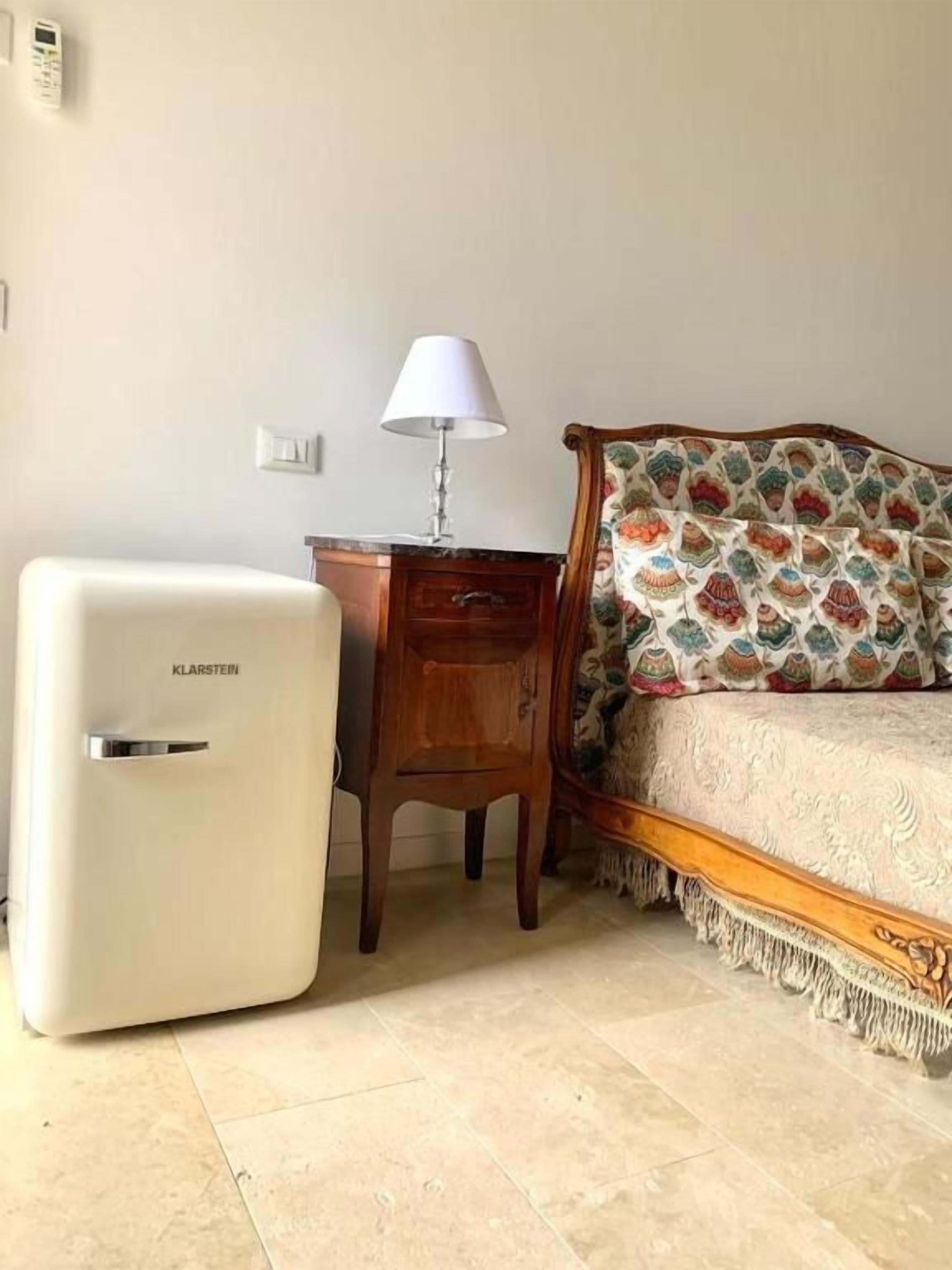 Un dormitorio con una cama, una mesita de noche, una lámpara y un electrodoméstico blanco.