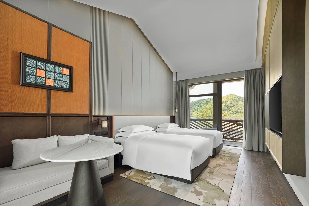 Habitación de hotel moderna con una cama grande, un sofá, una mesa de comedor y vistas a un paisaje montañoso.