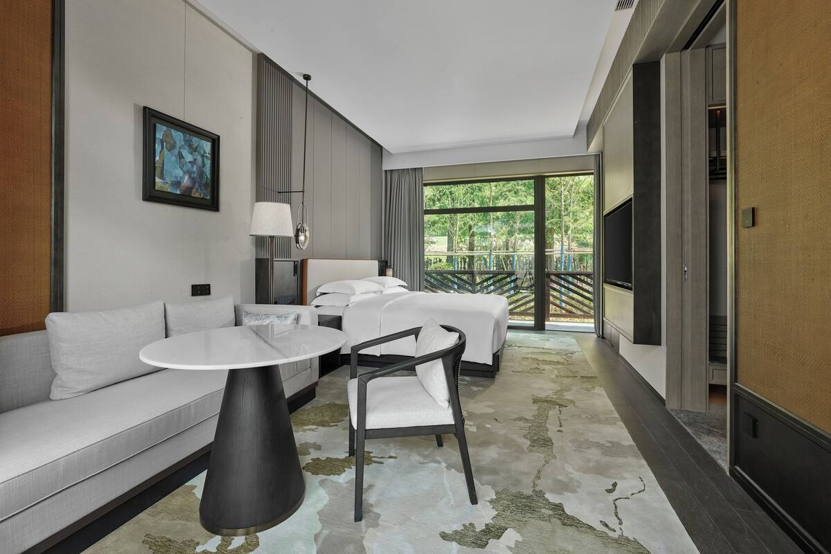 Habitación de hotel moderna con sofá, una mesita redonda, cama y balcón con vistas a árboles.