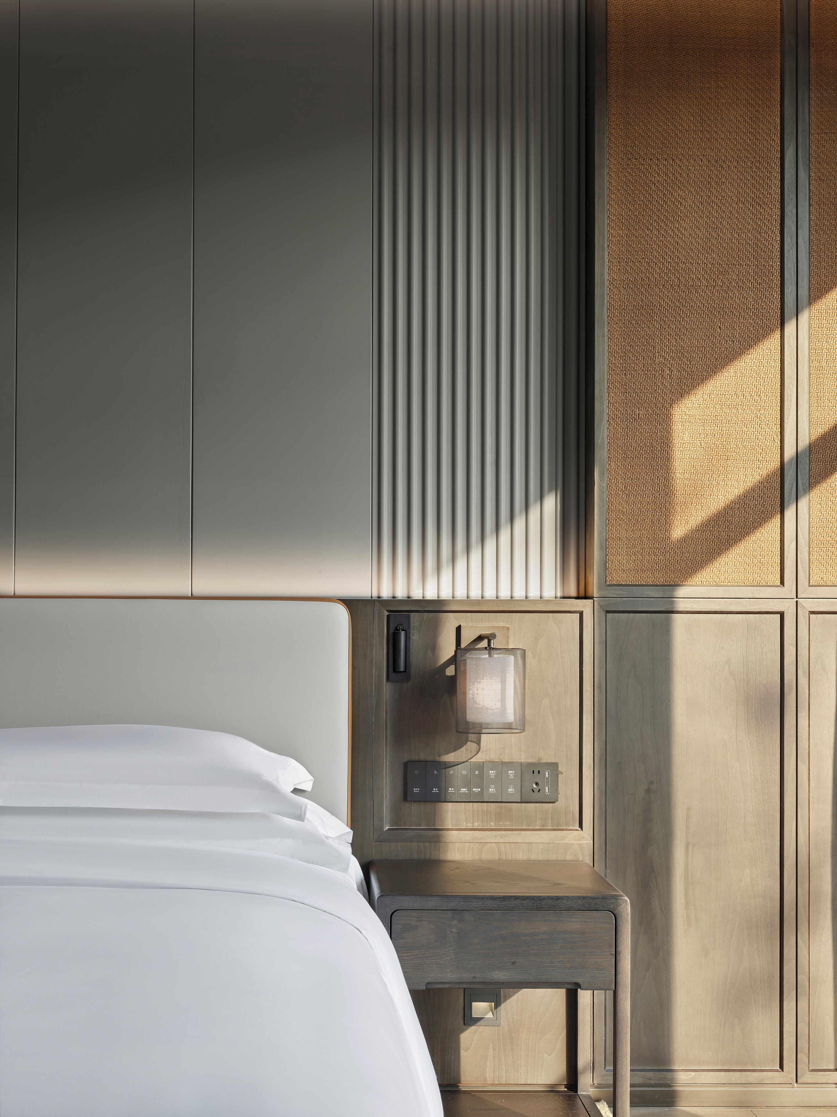 A hotel room with a bed, a bedside table with a lamp, and a power outlet.