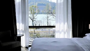 Ein Hotelzimmer mit einem großen Bett, einem Sessel und Blick auf einen See und Bäume durch transparente Vorhänge.