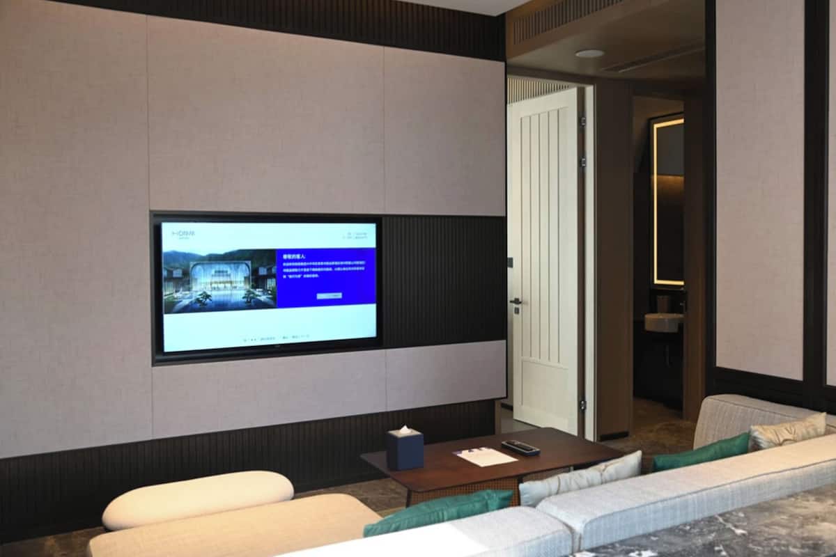 Habitación de hotel moderna con un televisor de pantalla plana grande montado en la pared, un sofá cómodo, una mesita y una puerta que da al baño.